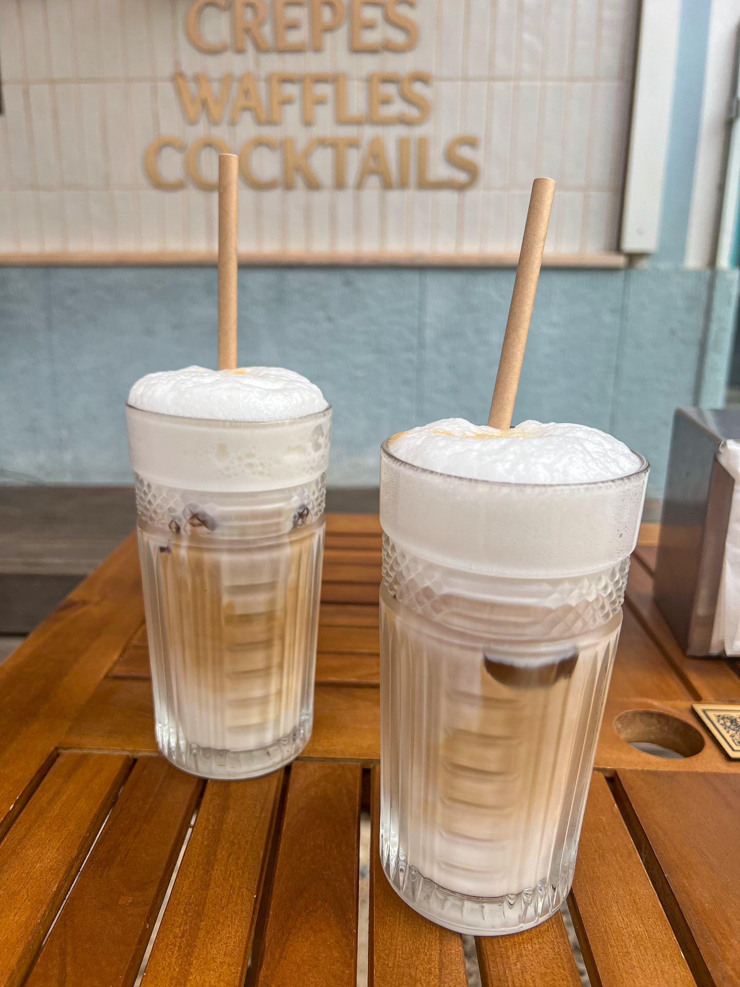 Two creamy iced drinks on a wooden café table with blurred sign in the background
