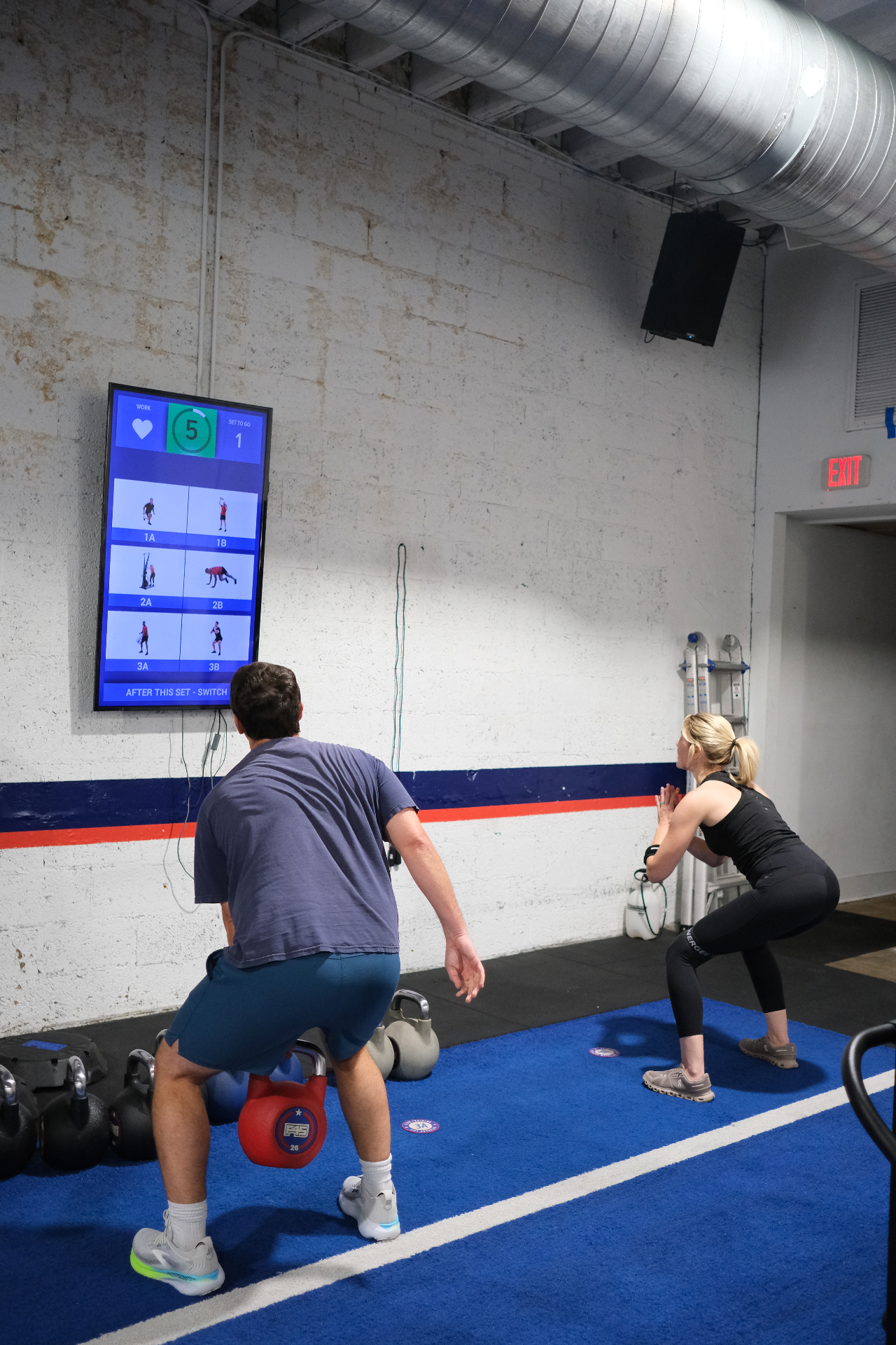 Two athletes mid-workout during a squat set in a blue-floor training studio