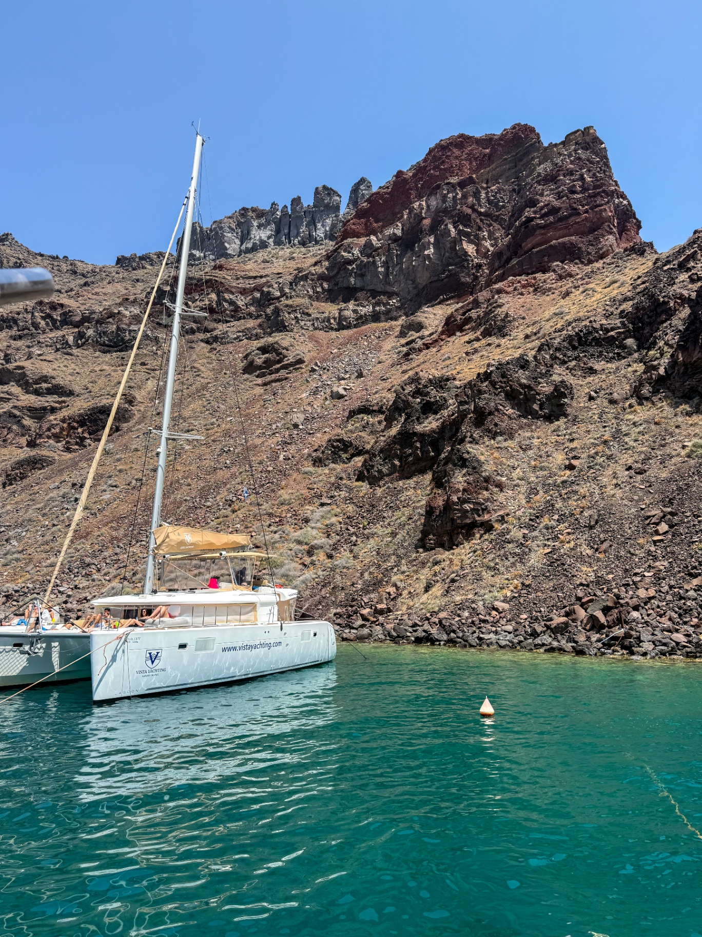 White catamaran anchored in clear turquoise water below a volcanic cliff