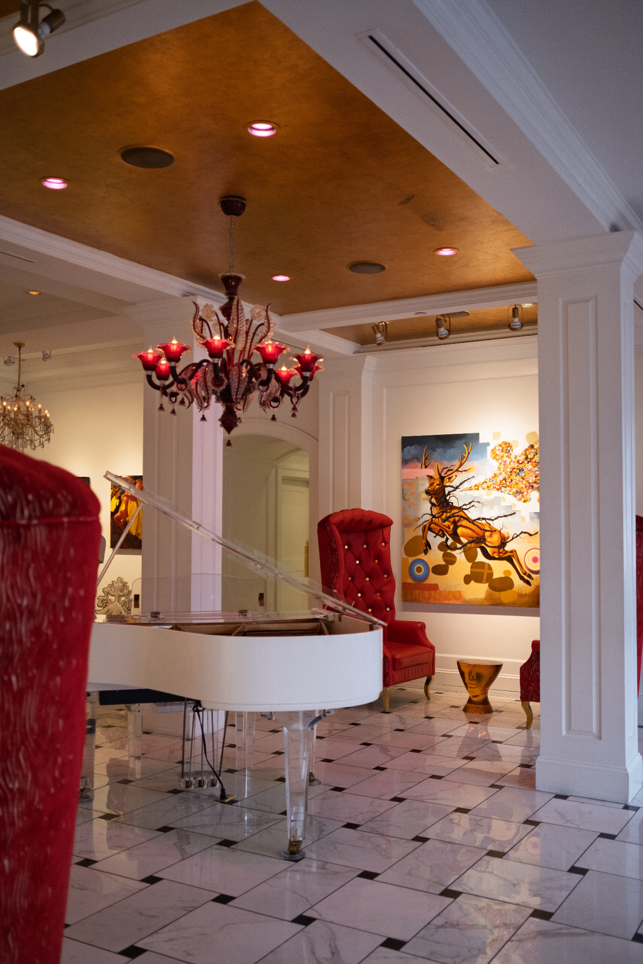 White grand piano under a red Murano chandelier in a Charleston gallery with bold contemporary paintings