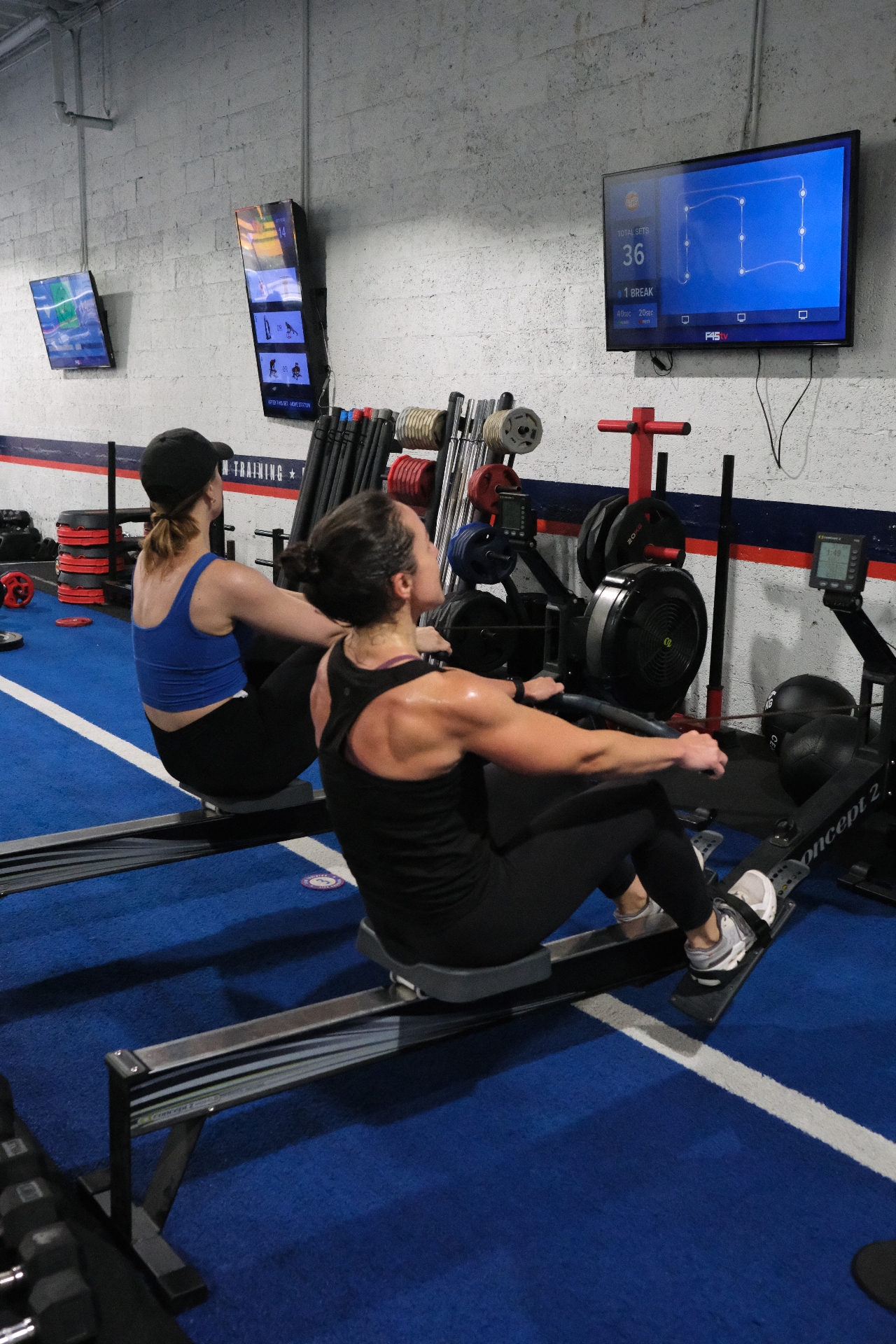 Two women rowing in sync in a strength studio surrounded by racks and screens